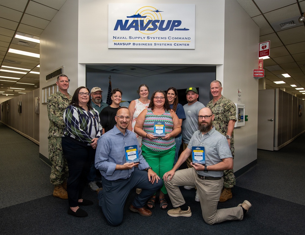 A group of approximately eleven people pose for a formal group photo in front of a NAVSUP (Naval Supply Systems Command) Business Systems Center sign. The group includes two uniformed Navy personnel and several civilians, with three individuals in the front holding small award plaques. This appears to be an award or recognition ceremony at a military facility.
