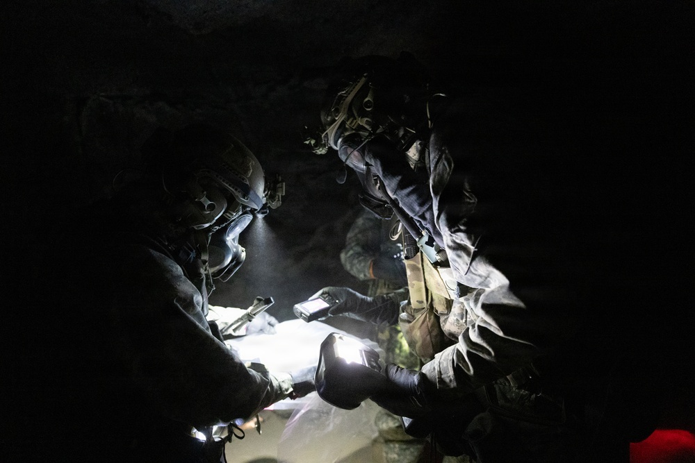 Two military personnel in full tactical gear are examining a map or document in a dark environment, illuminated by handheld flashlights and a helmet-mounted light. A third individual is partially visible in the background. The scene appears to depict a nighttime or low-light tactical planning or navigation activity.