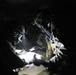 Two military personnel in full tactical gear are examining a map or document in a dark environment, illuminated by handheld flashlights and a helmet-mounted light. A third individual is partially visible in the background. The scene appears to depict a nighttime or low-light tactical planning or navigation activity.