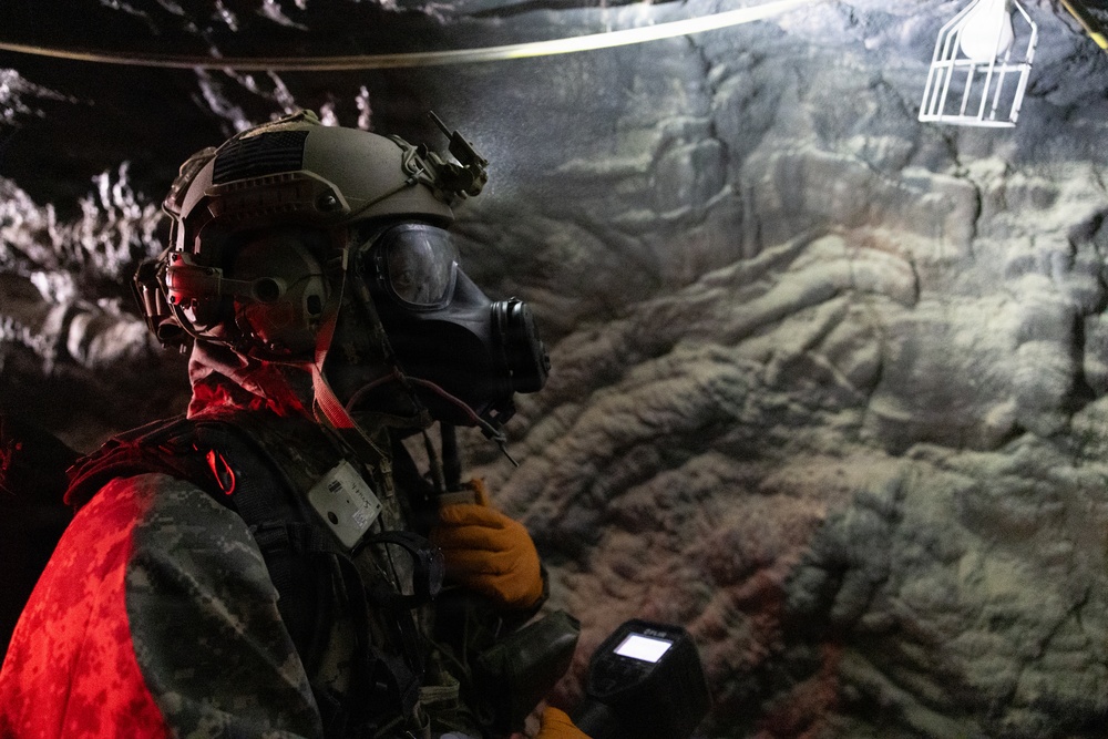 A military or tactical personnel is shown inside what appears to be a cave or underground tunnel environment. The individual is wearing full protective gear including a tactical helmet, gas mask, and camouflage uniform, and is holding a handheld device that appears to be a FLIR thermal imaging unit. The scene is dimly lit with a red light source illuminating the subject from the left side.