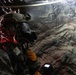 A military or tactical personnel is shown inside what appears to be a cave or underground tunnel environment. The individual is wearing full protective gear including a tactical helmet, gas mask, and camouflage uniform, and is holding a handheld device that appears to be a FLIR thermal imaging unit. The scene is dimly lit with a red light source illuminating the subject from the left side.