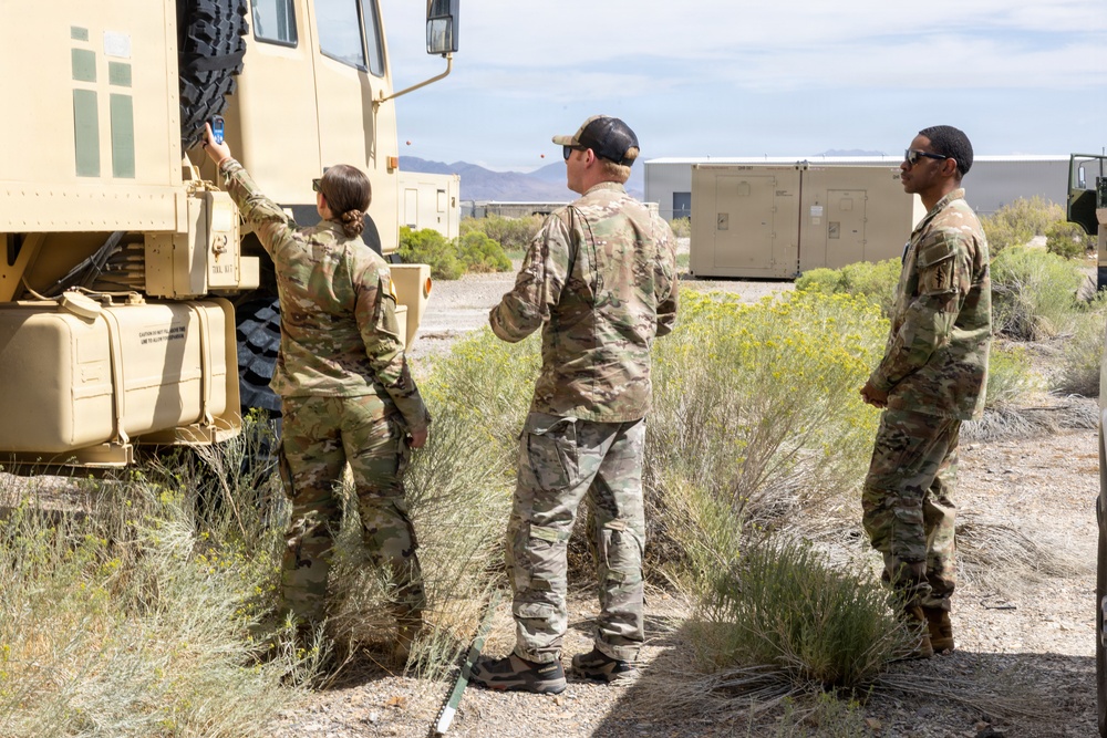 Three military personnel in OCP (Operational Camouflage Pattern) uniforms are gathered around a large tan military tactical vehicle in an arid desert environment. One soldier appears to be inspecting or interacting with the vehicle's side panel while the other two observe. The setting includes desert scrub vegetation, mountainous terrain in the background, and military container units nearby.