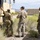 Three military personnel in OCP (Operational Camouflage Pattern) uniforms are gathered around a large tan military tactical vehicle in an arid desert environment. One soldier appears to be inspecting or interacting with the vehicle's side panel while the other two observe. The setting includes desert scrub vegetation, mountainous terrain in the background, and military container units nearby.