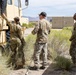 Three military personnel in OCP (Operational Camouflage Pattern) uniforms are gathered around a large tan military tactical vehicle in an arid desert environment. One soldier appears to be inspecting or interacting with the vehicle's side panel while the other two observe. The setting includes desert scrub vegetation, mountainous terrain in the background, and military container units nearby.