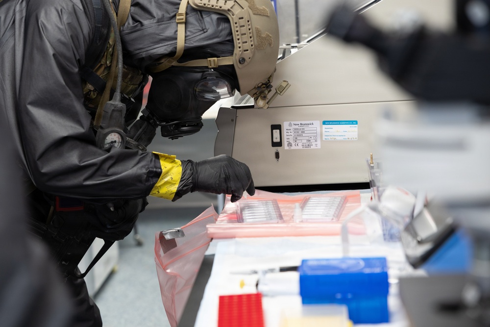 A person wearing full protective gear including a tactical helmet, gas mask, and black gloves with yellow tape markings is examining laboratory equipment and materials. The setting appears to be a scientific or medical laboratory environment, with various lab supplies visible on the work surface. The individual appears to be conducting an inspection or evidence collection activity.