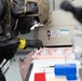 A person wearing full protective gear including a tactical helmet, gas mask, and black gloves with yellow tape markings is examining laboratory equipment and materials. The setting appears to be a scientific or medical laboratory environment, with various lab supplies visible on the work surface. The individual appears to be conducting an inspection or evidence collection activity.