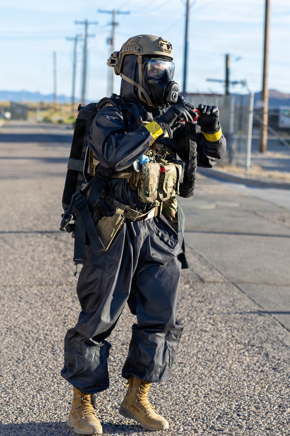 A military or law enforcement personnel is standing on a paved road wearing full CBRN (Chemical, Biological, Radiological, Nuclear) protective gear. The individual is equipped with a tactical helmet, full-face gas mask, protective overgarment suit, and multiple tactical pouches and accessories. The setting appears to be a desert or arid environment based on the terrain and utility poles visible in the background.