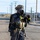 A military or law enforcement personnel is standing on a paved road wearing full CBRN (Chemical, Biological, Radiological, Nuclear) protective gear. The individual is equipped with a tactical helmet, full-face gas mask, protective overgarment suit, and multiple tactical pouches and accessories. The setting appears to be a desert or arid environment based on the terrain and utility poles visible in the background.