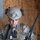 A U.S. Army soldier in full tactical gear is examining a small tan device with blue components, likely a training or electronic warfare tool. The soldier is wearing Operational Camouflage Pattern (OCP) uniform, a tactical helmet with NVG mount and communications headset, and a plate carrier vest with MOLLE attachments. The setting appears to be a training environment with an OSB/plywood wall in the background.