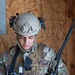 A U.S. Army soldier in full tactical gear is examining a small tan device with blue components, likely a training or electronic warfare tool. The soldier is wearing Operational Camouflage Pattern (OCP) uniform, a tactical helmet with NVG mount and communications headset, and a plate carrier vest with MOLLE attachments. The setting appears to be a training environment with an OSB/plywood wall in the background.