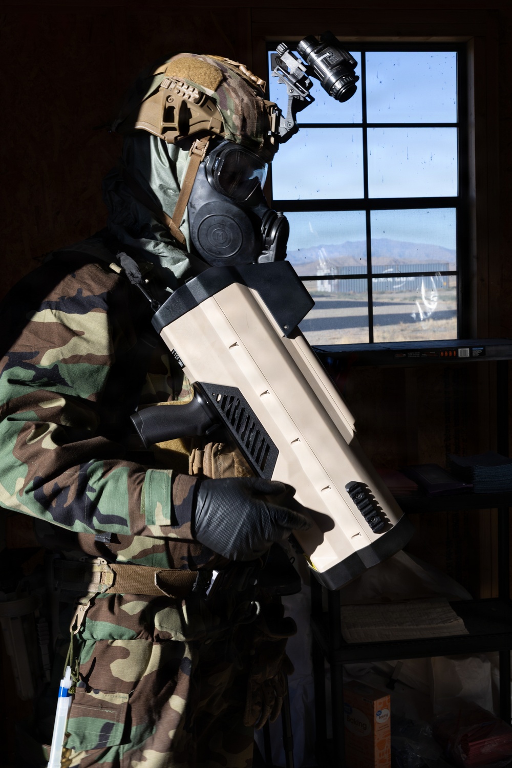 A person in full military/tactical gear is standing inside a building, holding a large tan-colored device that appears to be a counter-drone or directed-energy weapon system. The individual is wearing woodland camouflage, a gas mask, tactical helmet with night-vision device mount, and black gloves. The background shows a desert landscape visible through a multi-pane window.
