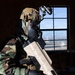 A person in full military/tactical gear is standing inside a building, holding a large tan-colored device that appears to be a counter-drone or directed-energy weapon system. The individual is wearing woodland camouflage, a gas mask, tactical helmet with night-vision device mount, and black gloves. The background shows a desert landscape visible through a multi-pane window.