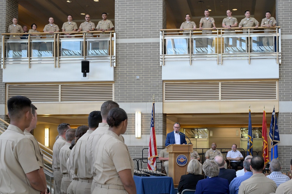 A formal military ceremony is taking place inside a modern building atrium, with a civilian speaker addressing an audience from a wooden podium bearing an official seal. Numerous uniformed military personnel in tan/khaki uniforms are observing from a second-floor balcony railing, while additional personnel and civilians are seated or standing on the ground floor. American and military branch flags are displayed prominently behind the speaker.