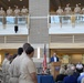 A formal military ceremony is taking place inside a modern building atrium, with a civilian speaker addressing an audience from a wooden podium bearing an official seal. Numerous uniformed military personnel in tan/khaki uniforms are observing from a second-floor balcony railing, while additional personnel and civilians are seated or standing on the ground floor. American and military branch flags are displayed prominently behind the speaker.
