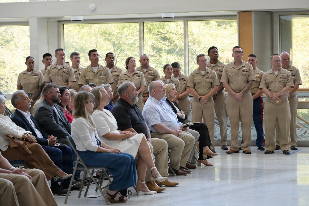 A formal military ceremony or event is taking place in a modern indoor facility with large windows. A group of uniformed U.S. Navy personnel (and at least one Marine) stand at attention in the background, while civilian guests and possibly family members are seated in folding chairs in the foreground. All attendees appear to be focused on something or someone outside the camera frame.