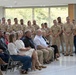 A formal military ceremony or event is taking place in a modern indoor facility with large windows. A group of uniformed U.S. Navy personnel (and at least one Marine) stand at attention in the background, while civilian guests and possibly family members are seated in folding chairs in the foreground. All attendees appear to be focused on something or someone outside the camera frame.
