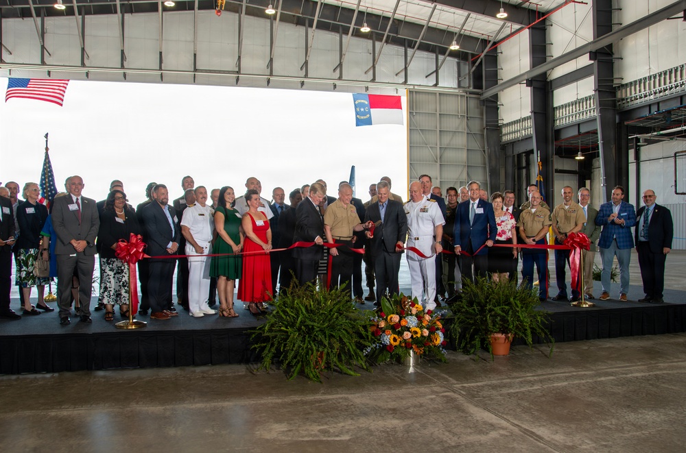 A formal ribbon-cutting ceremony is taking place inside a large industrial hangar facility. The event features a mixed group of military personnel in uniform and civilians on a raised stage, with an American flag and what appears to be a North Carolina state flag visible in the background. The ceremony appears to be a dedication or grand opening of a military or government facility.