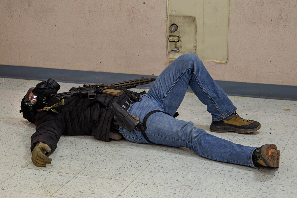 A person wearing tactical gear including a gas mask, body armor/vest, and gloves is lying on a tiled floor in what appears to be an indoor training facility. The individual is equipped with a rifle and a holstered sidearm, suggesting this is a law enforcement or military training exercise. The setting, including paint splatters on the walls, is consistent with a simulated training environment.