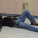 A person wearing tactical gear including a gas mask, body armor/vest, and gloves is lying on a tiled floor in what appears to be an indoor training facility. The individual is equipped with a rifle and a holstered sidearm, suggesting this is a law enforcement or military training exercise. The setting, including paint splatters on the walls, is consistent with a simulated training environment.