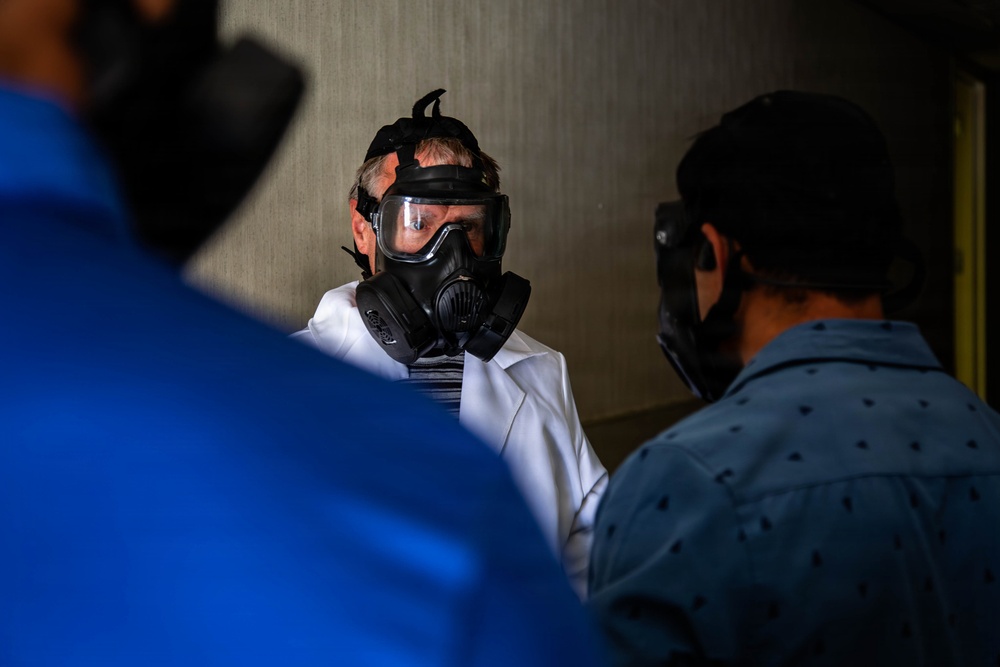 A person wearing a full-face gas mask and a white coat or shirt is engaged in conversation with at least two other individuals in what appears to be an indoor setting. The central figure's gas mask features dual filter canisters and a wide visor. The scene suggests some form of briefing, discussion, or demonstration involving protective respiratory equipment.