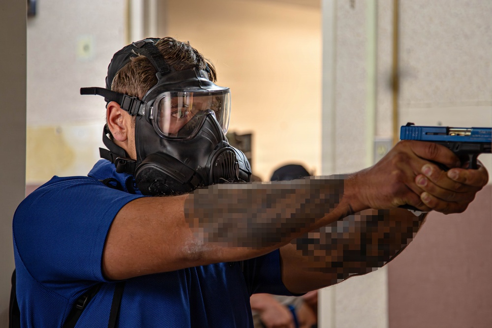 A person wearing a full-face gas mask/respirator and a blue polo shirt is aiming a blue-framed training pistol in what appears to be an indoor training facility. The individual has tattooed arms, with the tattoos partially pixelated in the image. The setting and equipment suggest a law enforcement or military training exercise.