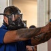 A person wearing a full-face gas mask/respirator and a blue polo shirt is aiming a blue-framed training pistol in what appears to be an indoor training facility. The individual has tattooed arms, with the tattoos partially pixelated in the image. The setting and equipment suggest a law enforcement or military training exercise.
