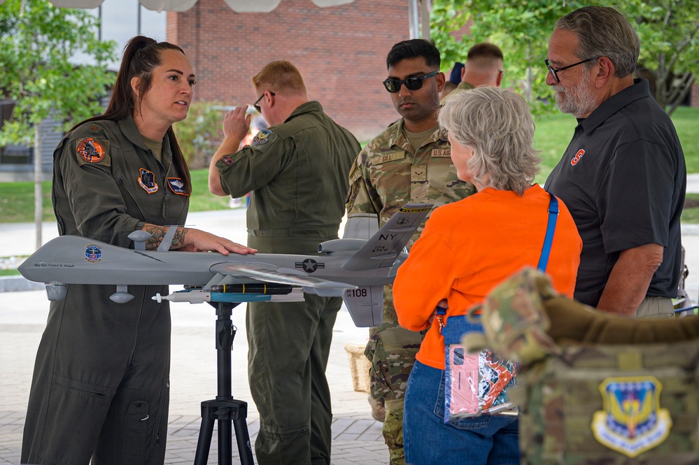 A female military aviator in an olive flight suit is presenting a scale model of a U.S. Air Force MQ-9 Reaper drone to civilian visitors at what appears to be a public outreach or community relations event. The model is mounted on a tripod stand and displays 'NY' markings and the number '108', suggesting it represents an aircraft from a New York Air National Guard unit. Several other military personnel in flight suits and camouflage uniforms are present in the background.