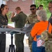 A female military aviator in an olive flight suit is presenting a scale model of a U.S. Air Force MQ-9 Reaper drone to civilian visitors at what appears to be a public outreach or community relations event. The model is mounted on a tripod stand and displays 'NY' markings and the number '108', suggesting it represents an aircraft from a New York Air National Guard unit. Several other military personnel in flight suits and camouflage uniforms are present in the background.