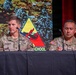 Two U.S. Army officers are seated at a panel table during what appears to be a formal military briefing or symposium. The officer on the left is identified by his name tape as 'ZIEGELHOFER' and holds the rank of Colonel (silver eagle insignia visible), while the officer on the right is partially identified as 'PULID-' (name partially obscured). A military unit insignia — appearing to be an Armor School patch — is projected on the screen behind them.