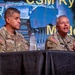 Two U.S. Army soldiers in camouflage uniforms are seated at a panel table during what appears to be a military conference or symposium. The soldier on the right is speaking into a microphone, while the one on the left listens. A projected screen behind them displays text including 'CSM Ryan' and 'Moderat[or]'.