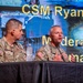 Two U.S. Army soldiers in camouflage uniforms are seated at a panel table during a military conference or briefing event. The soldier on the right is speaking into a microphone while the one on the left listens. A projected screen behind them displays text including 'CSM Ryan Dil...' and 'Moderator' and 'mande...' suggesting a formal panel discussion.