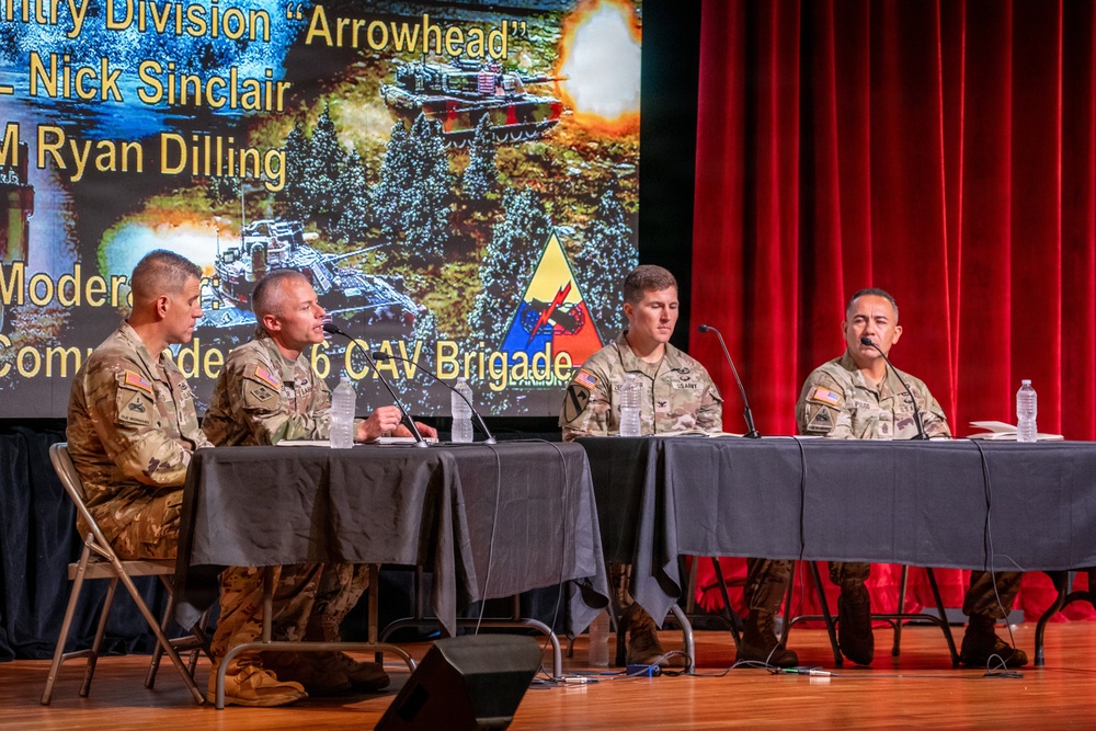 Four U.S. Army soldiers in camouflage uniforms are seated at a panel table on a stage, participating in a military briefing or symposium. The presentation slide behind them references the 'Arrowhead' Infantry Division, names Nick Sinclair and Ryan Dilling, and mentions a '6 CAV Brigade' commander role. A military unit flag with a triangle insignia is visible on the table.