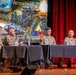 Four U.S. Army soldiers in camouflage uniforms are seated at a panel table on a stage, participating in a military briefing or symposium. The presentation slide behind them references the 'Arrowhead' Infantry Division, names Nick Sinclair and Ryan Dilling, and mentions a '6 CAV Brigade' commander role. A military unit flag with a triangle insignia is visible on the table.