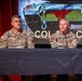 Two U.S. Army personnel in camouflage uniforms are seated at a draped panel table, apparently participating in a briefing or press conference. A projection screen behind them displays what appears to be a military armored vehicle and text reading 'COL' and 'Cann...' along with partial name identifiers. Name tapes on their uniforms read 'DILLING' and 'SINCLAIR'.