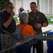 A U.S. Air Force pilot in an olive flight suit is speaking with two civilians at what appears to be a military outreach or public affairs event. Scale models of unmanned aerial vehicles (UAVs/drones) are displayed on a blue-draped table in the foreground. The setting appears to be an outdoor venue with a grass field visible in the background.
