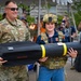 A military service member in OCP camouflage uniform and a young person wearing military gear (helmet, vest) pose together holding a training/display model of an AGM-114 Hellfire missile labeled 'M34' at what appears to be a public military outreach or recruitment event. The child is dressed in military equipment for a photo opportunity. The setting appears to be an outdoor public event with spectators visible in the background.