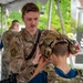 A U.S. Air Force servicemember named McCrink, wearing OCP (Operational Camouflage Pattern) uniform, is fitting a tactical helmet with ear protection onto a young person at what appears to be a public military outreach or recruitment event. The setting appears to be an outdoor venue, possibly a university campus, with civilians in orange shirts visible in the background. This appears to be a community engagement or demonstration event.
