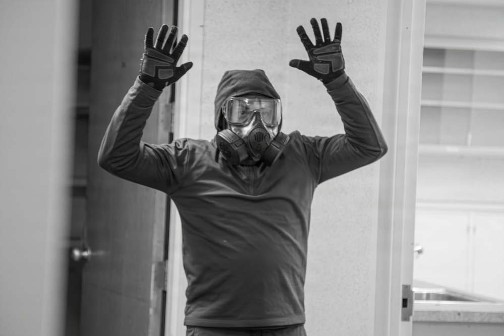A person wearing full protective gear including a respirator mask, large goggles, a hood, and tactical gloves stands with both hands raised in an indoor setting. The black-and-white photograph captures the individual in what appears to be a surrender or 'hands up' pose. The setting appears to be a residential or office interior.