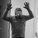 A person wearing full protective gear including a respirator mask, large goggles, a hood, and tactical gloves stands with both hands raised in an indoor setting. The black-and-white photograph captures the individual in what appears to be a surrender or 'hands up' pose. The setting appears to be a residential or office interior.