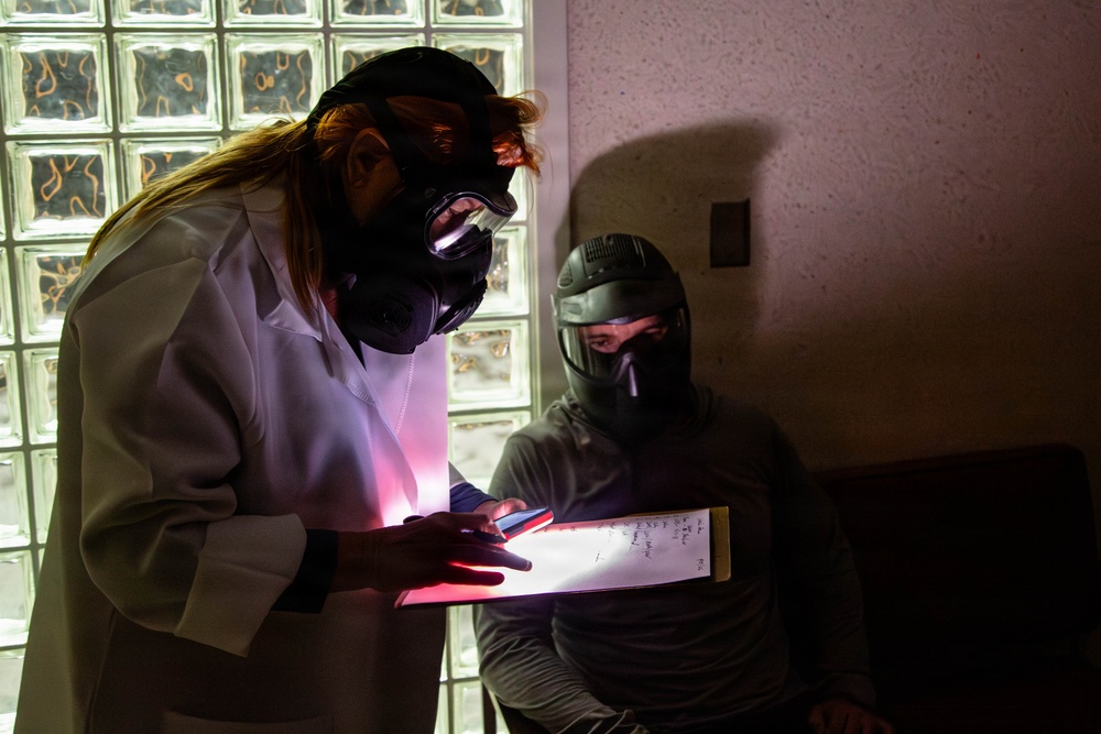 Two individuals wearing protective respiratory masks and gear are examining an illuminated clipboard or document in a dimly lit room. One person wears a white lab coat and full-face gas mask with long hair visible, while the other wears a dark tactical-style helmet and mask. The scene appears to be a staged or theatrical setting, possibly a film production, escape room, or training exercise.
