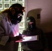 Two individuals wearing protective respiratory masks and gear are examining an illuminated clipboard or document in a dimly lit room. One person wears a white lab coat and full-face gas mask with long hair visible, while the other wears a dark tactical-style helmet and mask. The scene appears to be a staged or theatrical setting, possibly a film production, escape room, or training exercise.