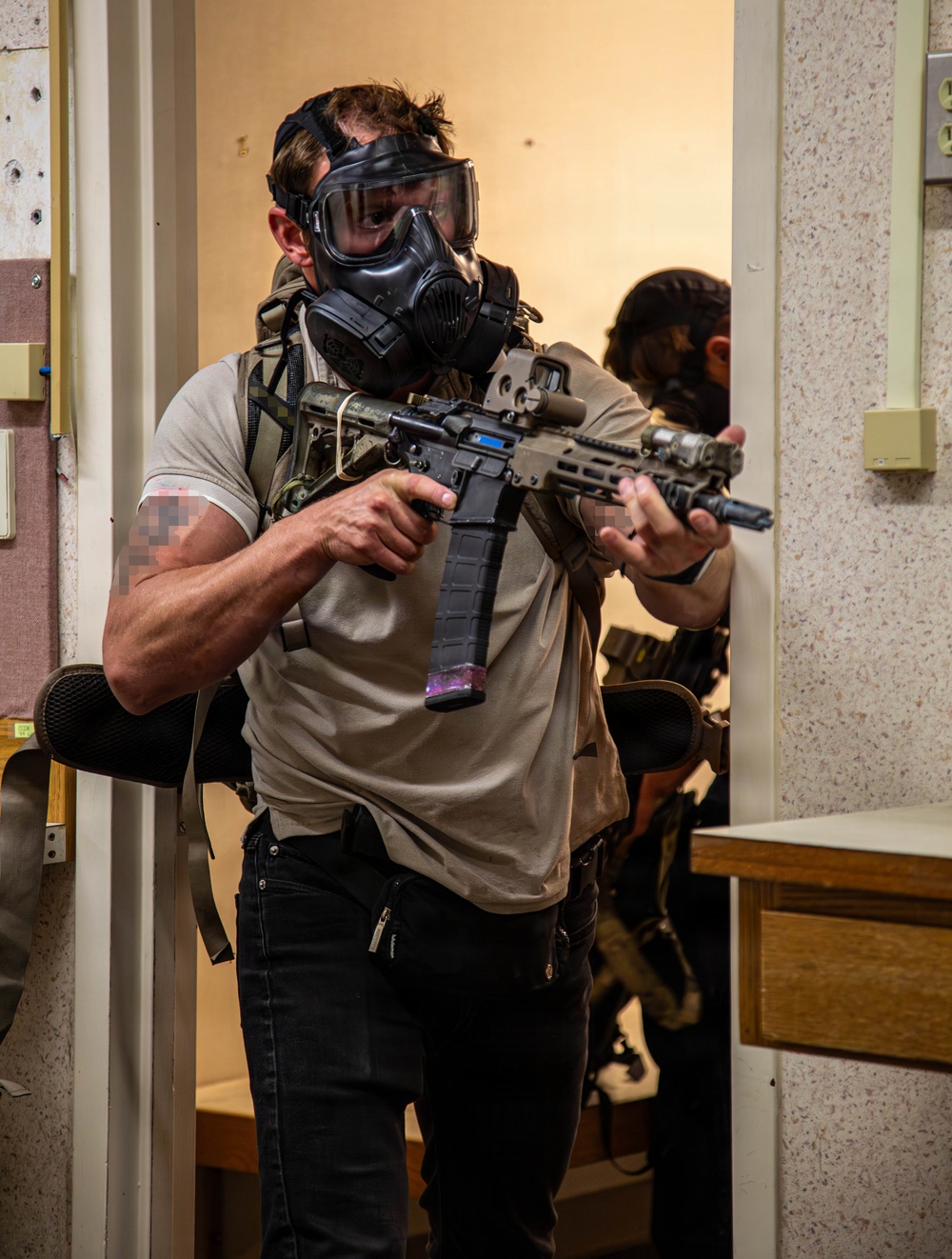 Two individuals wearing gas masks and tactical gear are conducting what appears to be a close-quarters building clearance drill or training exercise indoors. The primary subject in the foreground is holding a short-barreled AR-style rifle with optics and accessories, wearing civilian clothing with a tactical chest rig. A second individual is visible in the background performing a similar action.