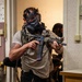 Two individuals wearing gas masks and tactical gear are conducting what appears to be a close-quarters building clearance drill or training exercise indoors. The primary subject in the foreground is holding a short-barreled AR-style rifle with optics and accessories, wearing civilian clothing with a tactical chest rig. A second individual is visible in the background performing a similar action.