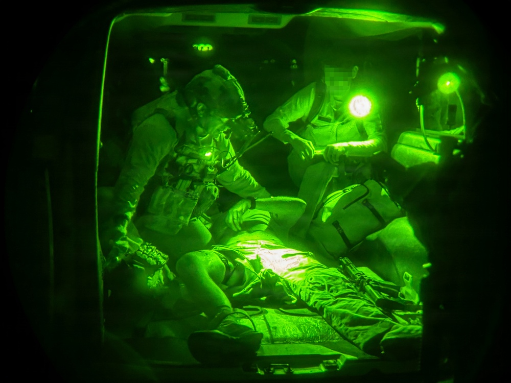 A night-vision photograph showing military medical personnel attending to a casualty on a stretcher, likely inside a helicopter or aircraft. The image is captured through a night-vision device, rendering everything in characteristic green monochrome. At least two personnel wearing tactical gear and helmets are providing medical care to a person lying supine on a litter.