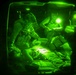 A night-vision photograph showing military medical personnel attending to a casualty on a stretcher, likely inside a helicopter or aircraft. The image is captured through a night-vision device, rendering everything in characteristic green monochrome. At least two personnel wearing tactical gear and helmets are providing medical care to a person lying supine on a litter.
