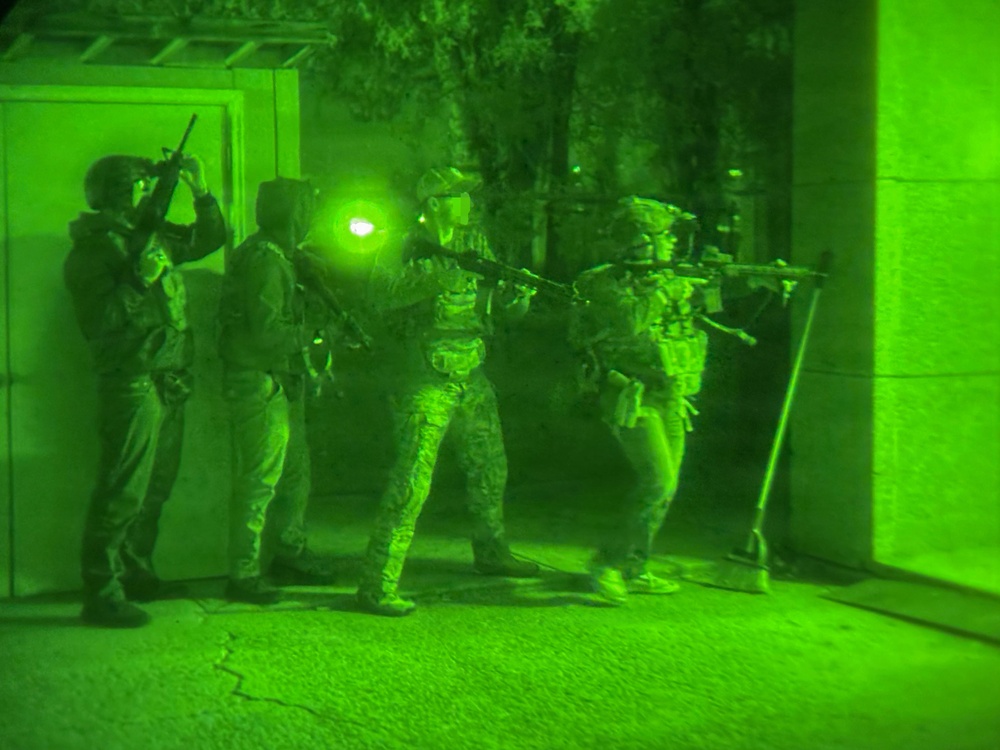 A night-vision captured image showing a group of armed military or law enforcement personnel conducting a tactical entry or building clearance operation. The image is rendered entirely in green monochrome tones characteristic of night-vision optics. Personnel are in full tactical gear and appear to be stacking near a doorway or entry point.