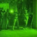 A night-vision captured image showing a group of armed military or law enforcement personnel conducting a tactical entry or building clearance operation. The image is rendered entirely in green monochrome tones characteristic of night-vision optics. Personnel are in full tactical gear and appear to be stacking near a doorway or entry point.