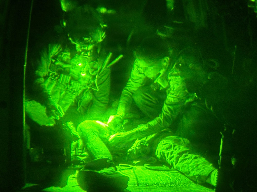 A night-vision captured image showing military personnel inside what appears to be a helicopter or aircraft interior. The green monochromatic illumination is characteristic of night-vision goggle (NVG) photography. Personnel appear to be attending to a seated individual, possibly in a medical or casualty evacuation context.