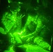 A night-vision captured image showing military personnel inside what appears to be a helicopter or aircraft interior. The green monochromatic illumination is characteristic of night-vision goggle (NVG) photography. Personnel appear to be attending to a seated individual, possibly in a medical or casualty evacuation context.