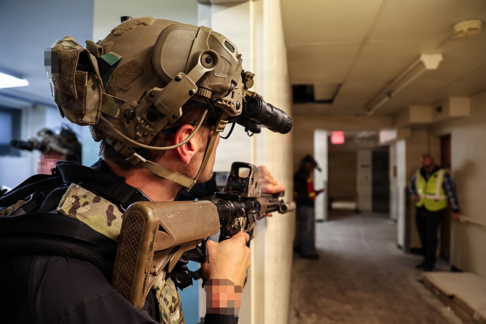 A heavily equipped tactical operator in full combat gear is conducting what appears to be a building clearance drill or training exercise in an indoor corridor. The operator is wearing a multicam helmet with night vision mount and aiming a rifle around a corner. Observers in high-visibility vests visible in the background suggest this is a supervised training or exercise scenario.